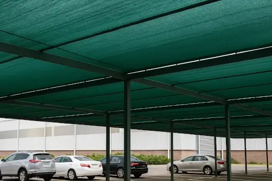 Estructura metálica de estacionamiento cubierta con polisombra verde que brinda protección solar a vehículos y maquinaria.